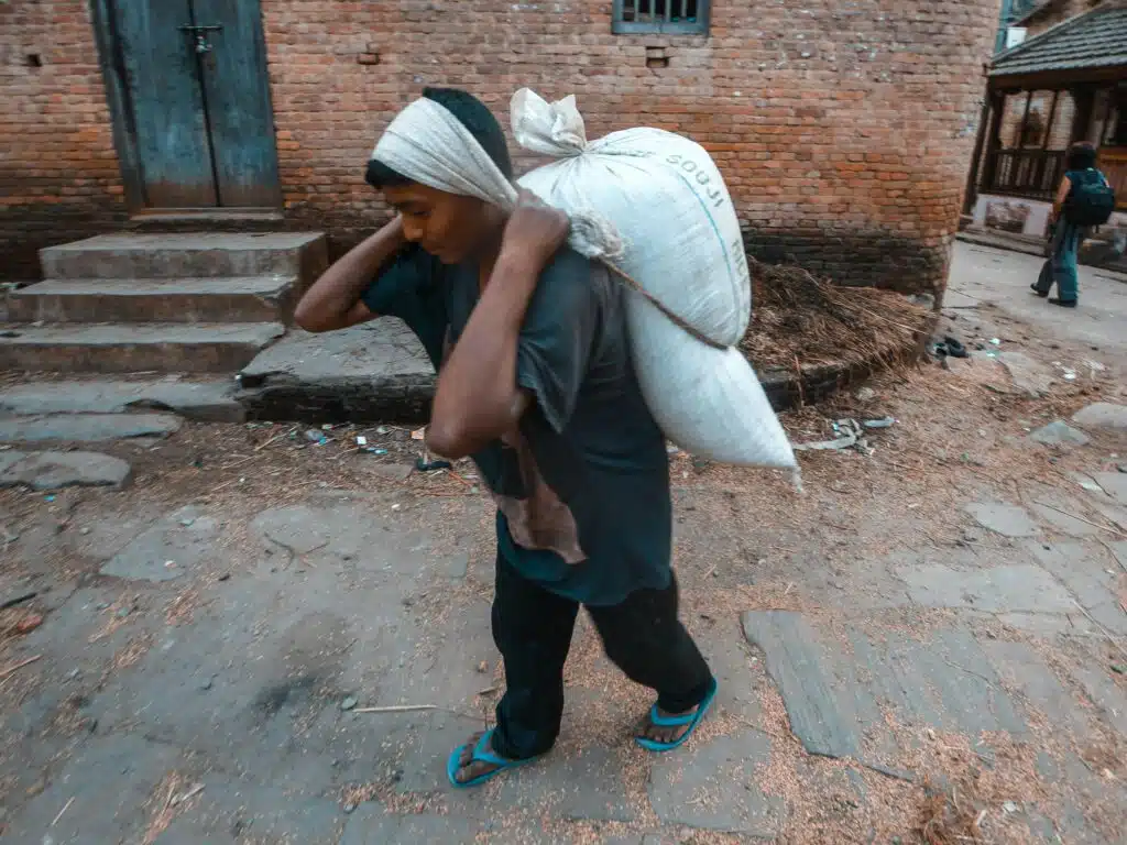 An adult man engaged in manual labor, carrying a heavy sack in an outdoor urban setting.