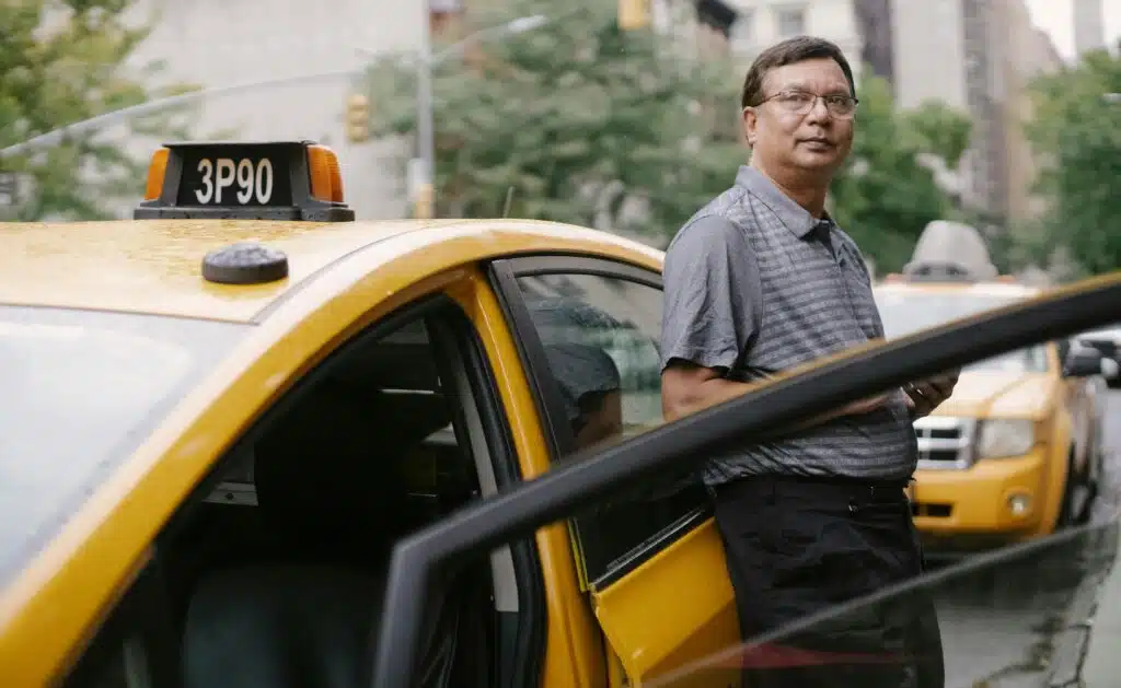 Asian taxi driver standing beside a yellow cab in the city with an open door.