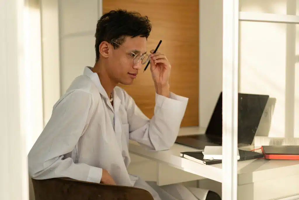 A young man wearing a white thobe sits indoors, pondering at a desk with a laptop and notes.