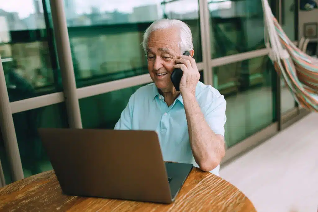 Elderly man in a blue shirt talking on phone while using laptop indoors. Ideal for remote work concepts.