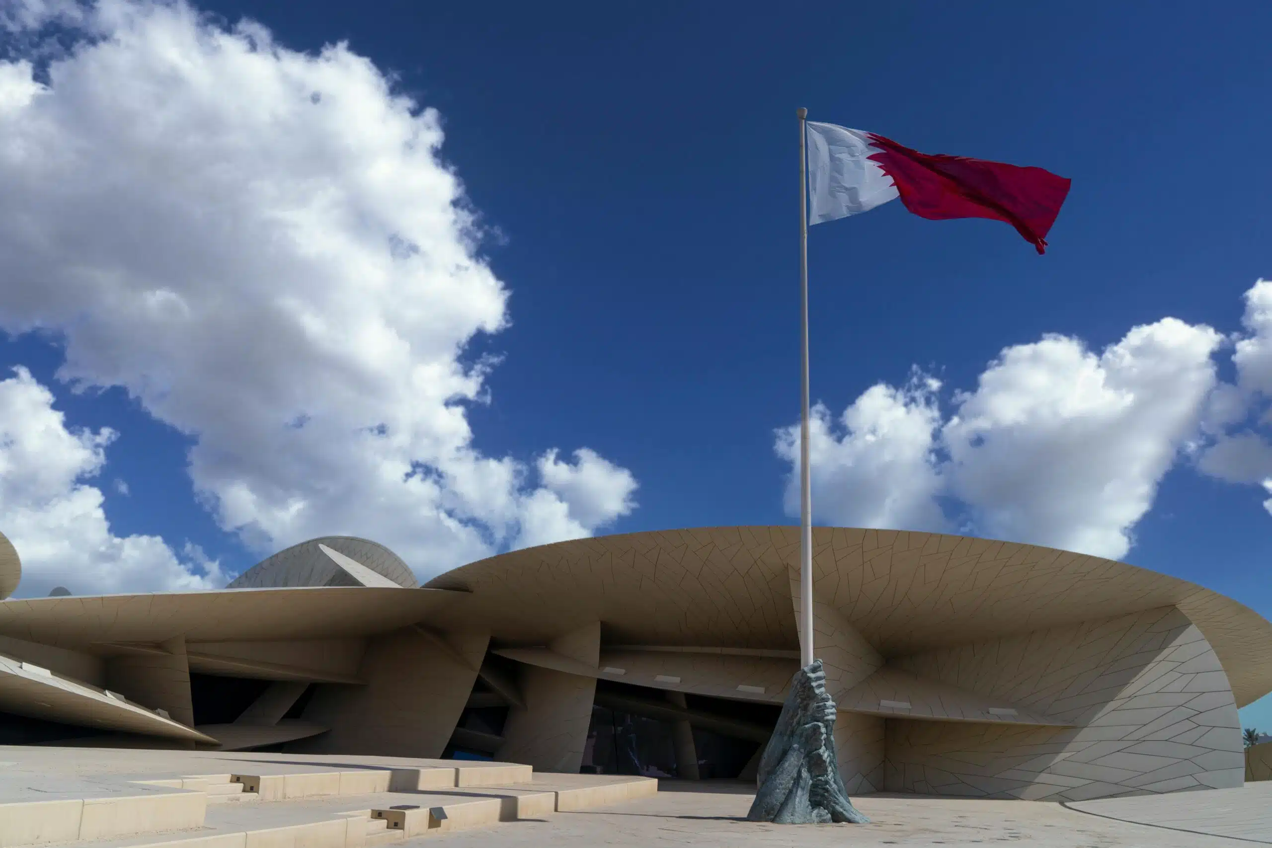 Qatar National Museum and flag against a vibrant blue sky with clouds in Doha.