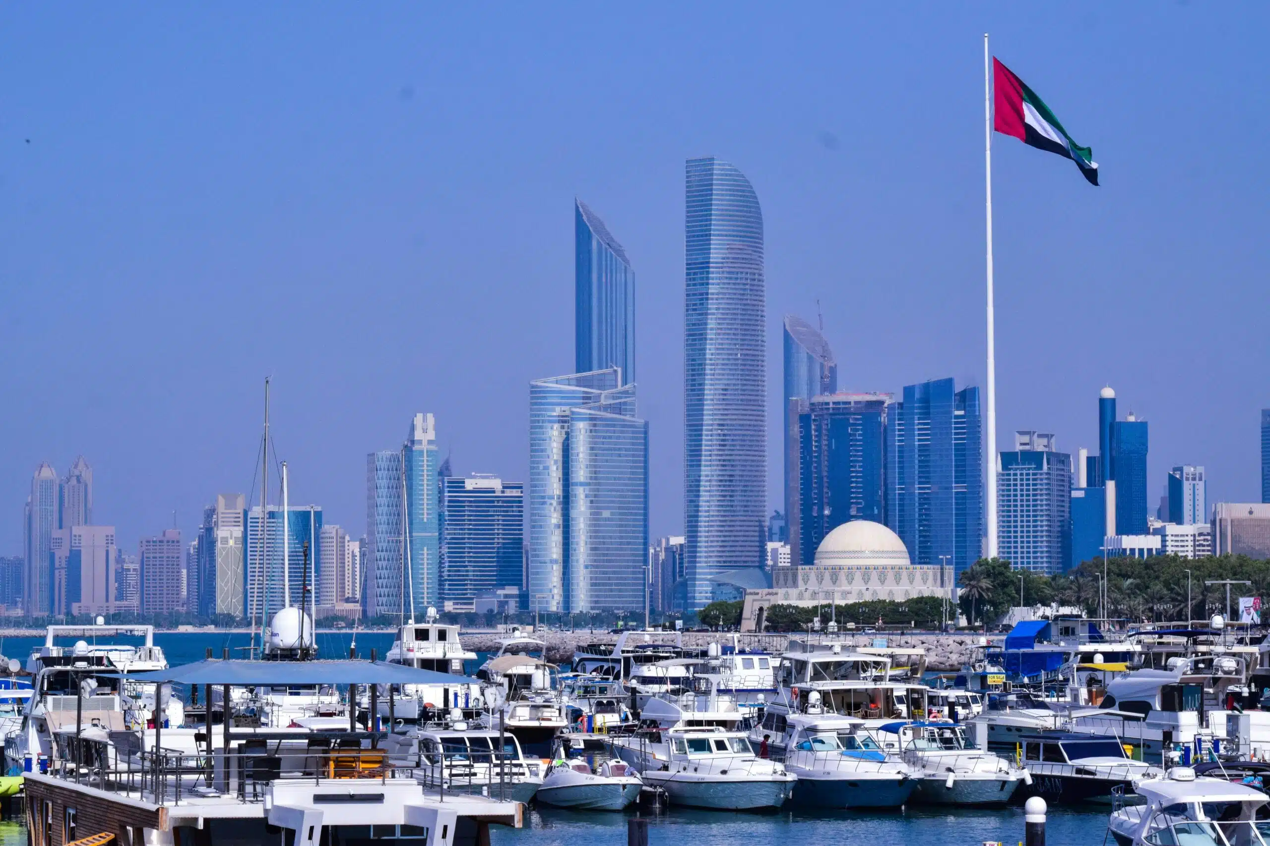 A scenic view of Abu Dhabi marina with modern skyscrapers and a flag in the foreground.